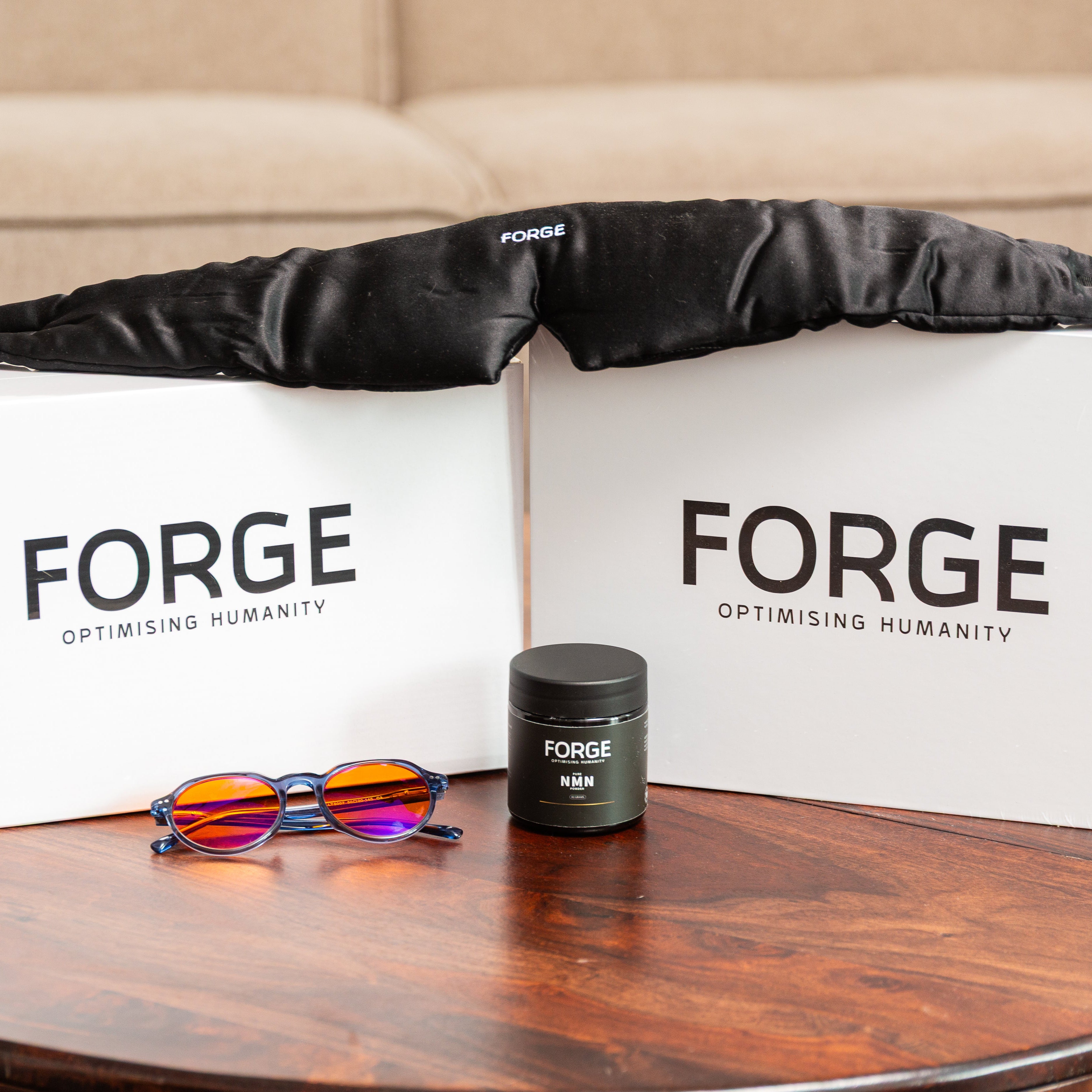 FORGE branded items including a black silk sleep mask, blue light blocking glasses, NMN supplement, boxes containing red light therapy devices on a wooden table.