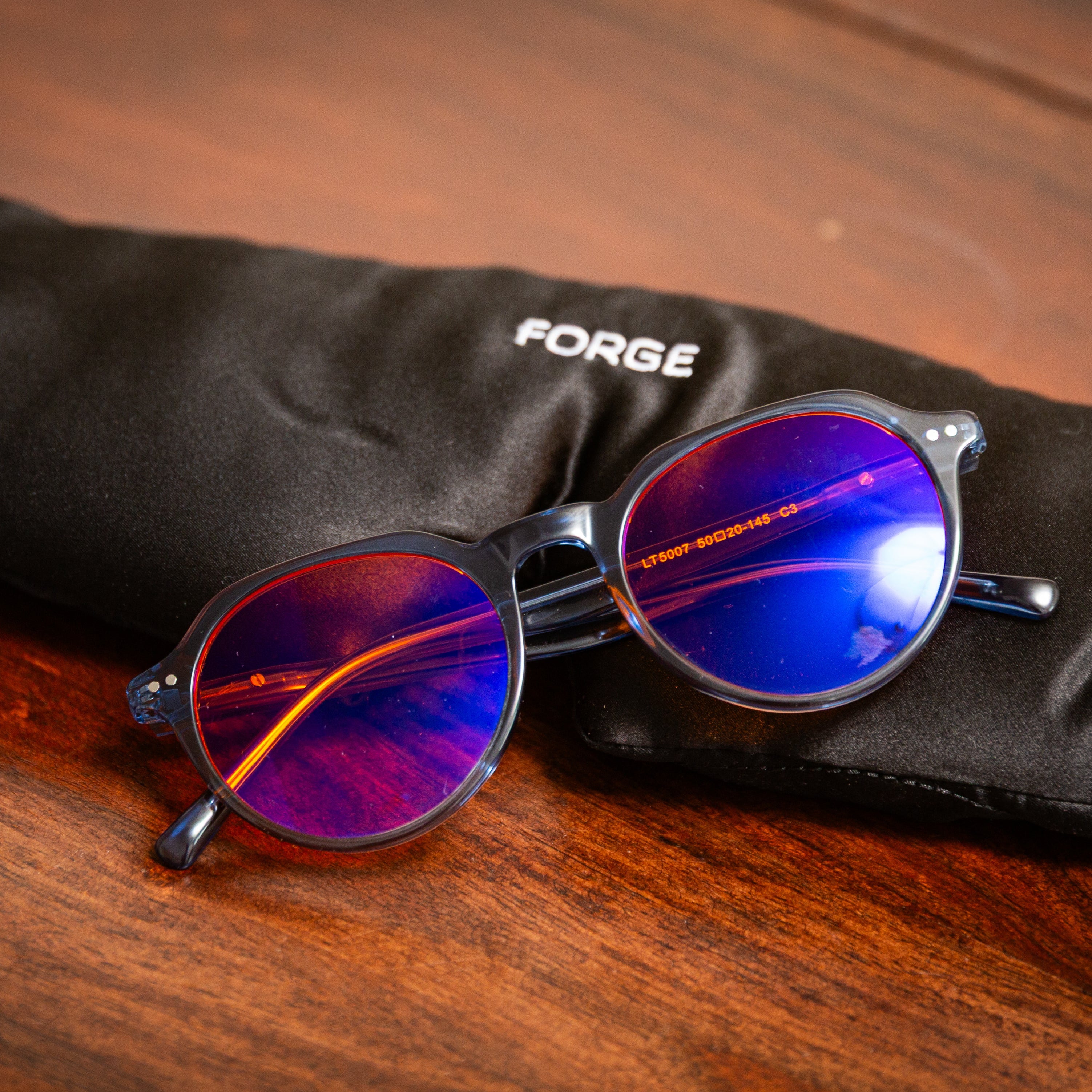 Round glasses with orange blue light blocking reflective lenses on a black 'FORGE' branded silk sleep mask on a wooden surface
