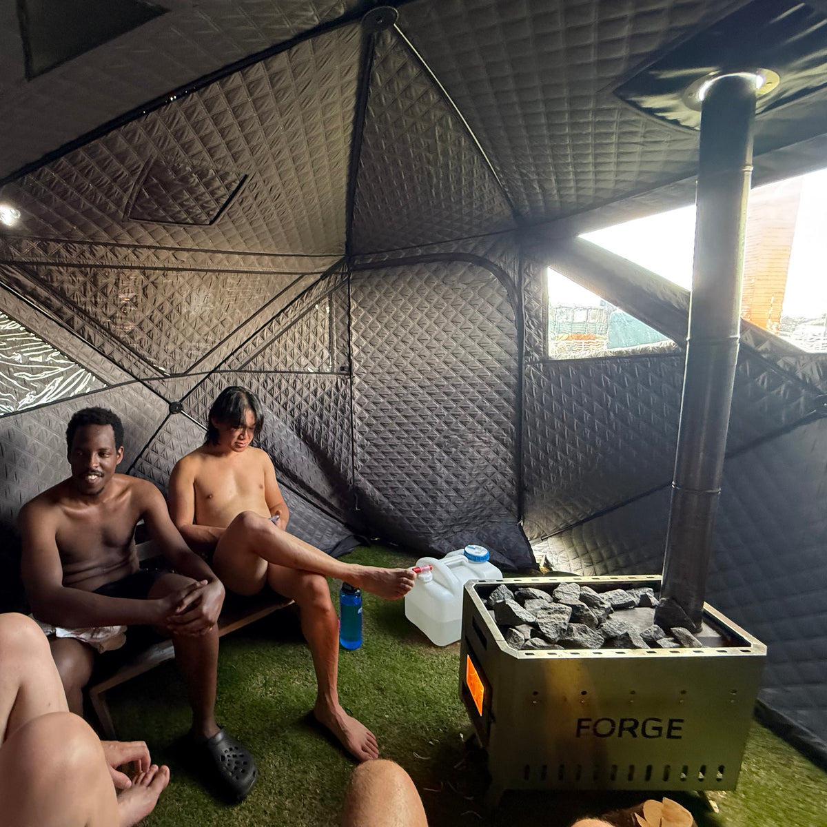 Three people sitting inside a heated Forge sauna tent with a wood-burning stove and artificial grass flooring.