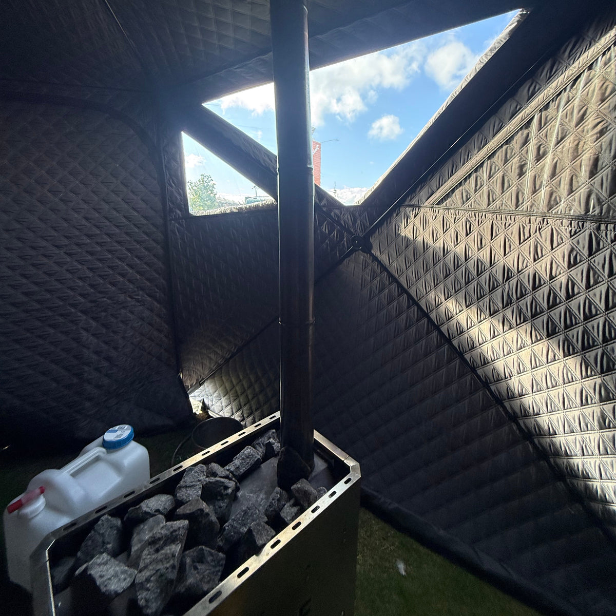 A dark metal wood-burning sauna stove with a chimney pipe rising up inside a quilted outdoor sauna tent, filled with charcoal blocks. A white plastic jerrycan is visible on the left, with a window showing blue sky and grass outside.