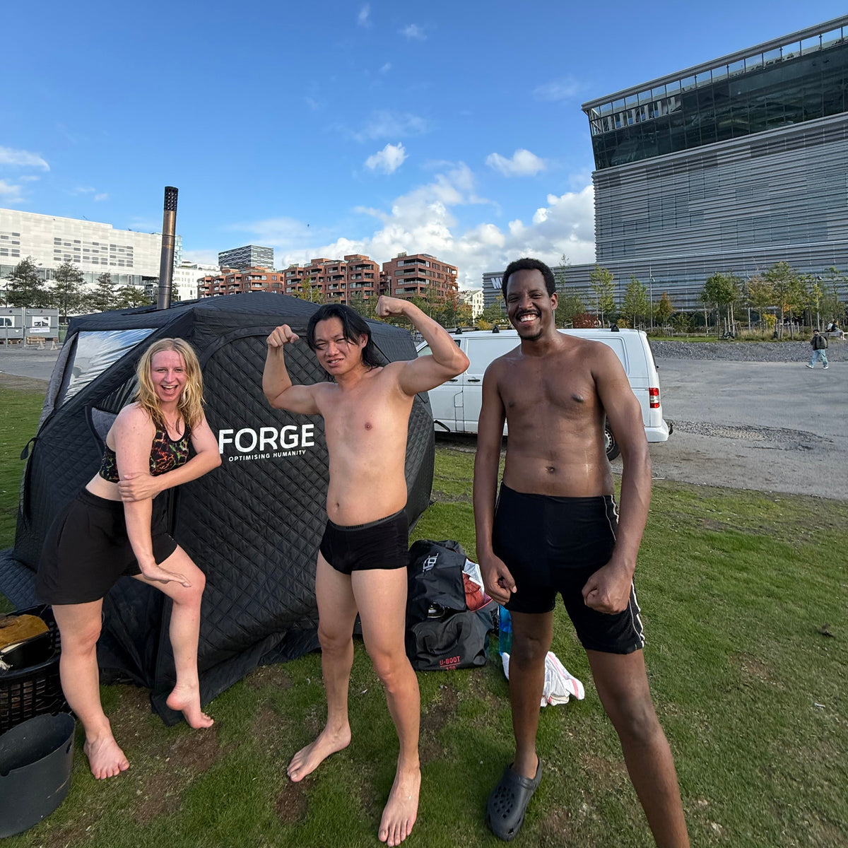 Three people posing outdoors in front of a black inflatable Badstutelt with the brand name FORGE visible on the side, set on a grassy lawn under a blue sky.