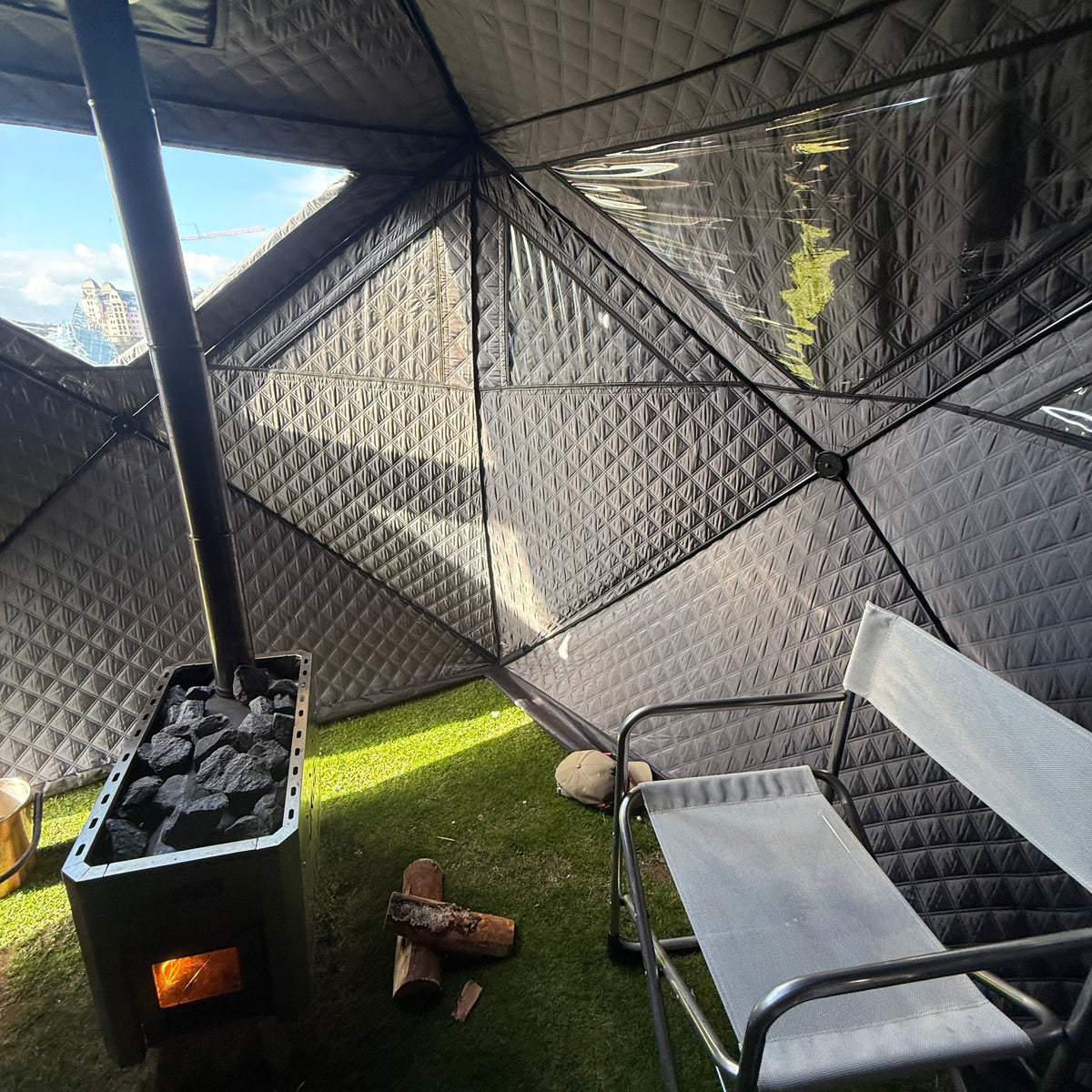 An interior view of a geodesic dome badstutelt with a black wood-burning sauna oven  stove, a white camping chair, and green grass flooring.